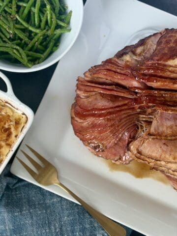 cooked spiral ham on white plate white bowl of green beans baking dish of au gratin potatoes