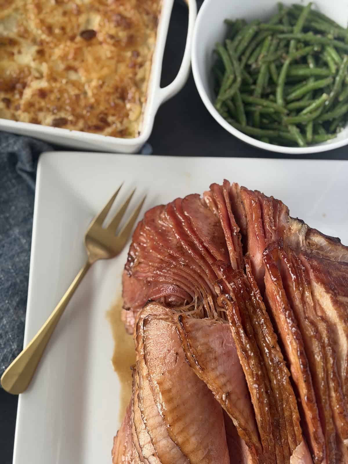 spiral ham on white plate gold serving fork baked au gratin potatoes bowl of green beans
