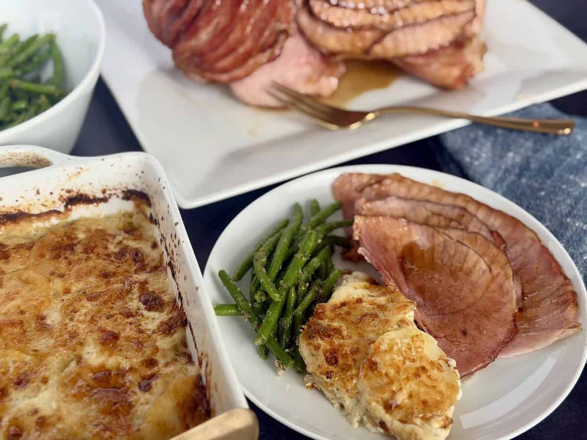 sliced ham au gratin potatoes and green beans on white plate next to serving dishes and blue napkin