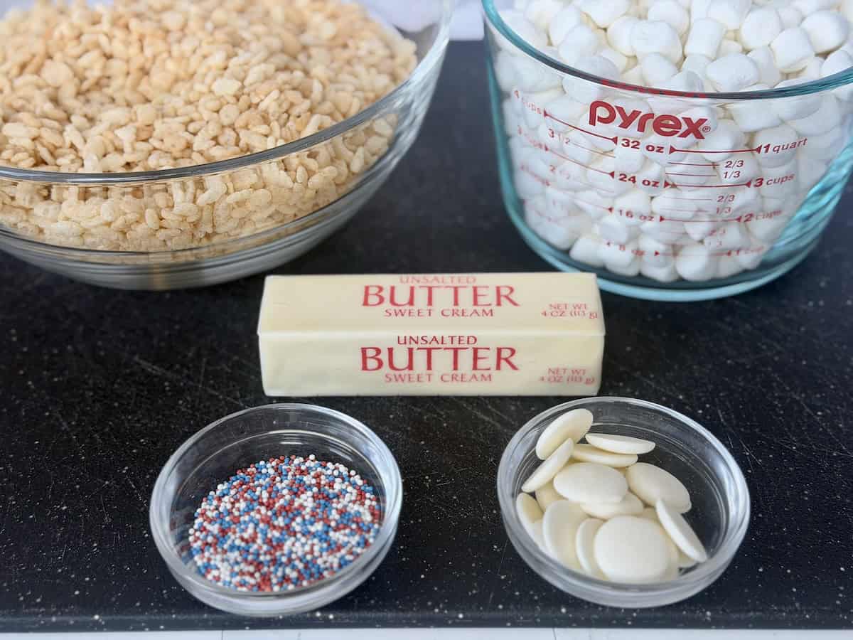 bowl of rice krispies cereal mini marshmallows butter sprinkles and white chocolate on black cutting board 