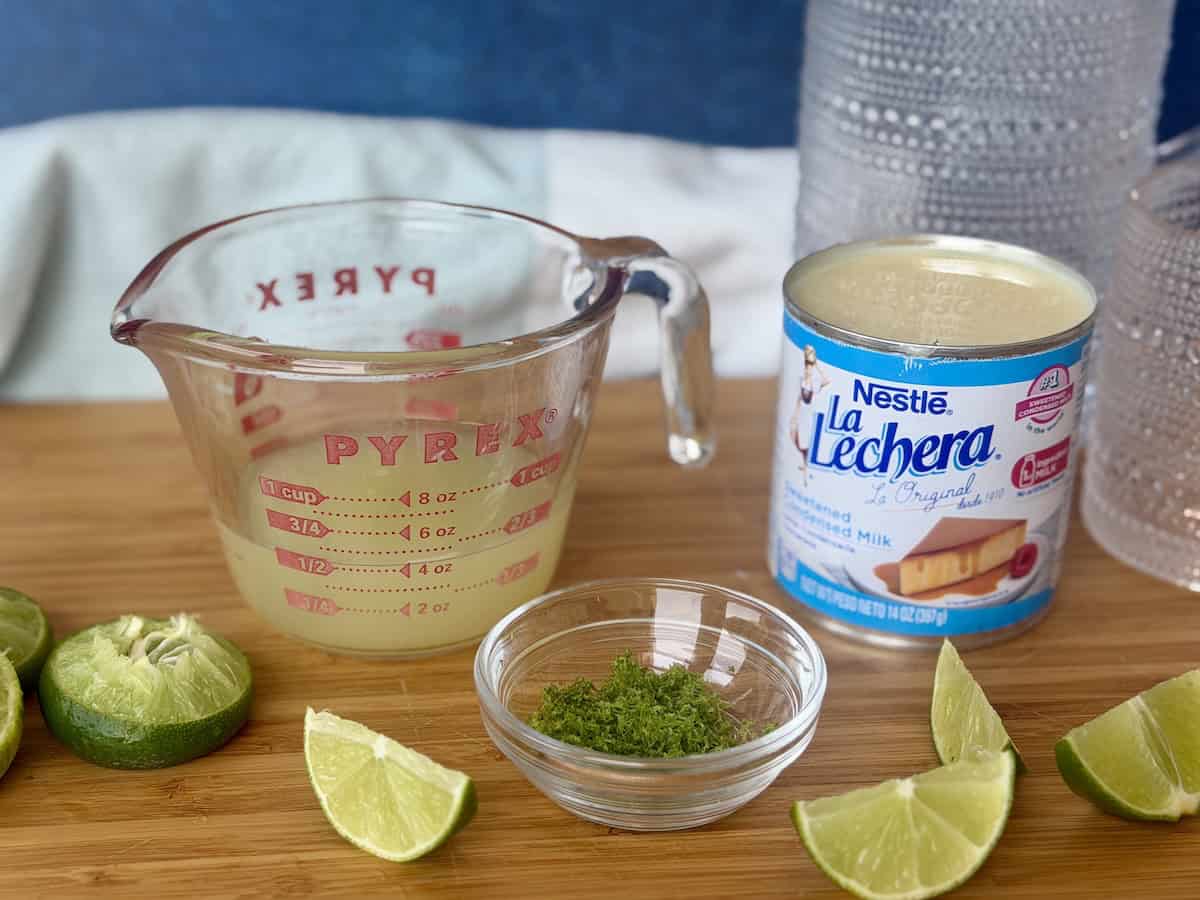 squeezed lime juice in measuring cup lime zest in glass bowl opened can of sweetened condensed milk all on wood cutting board