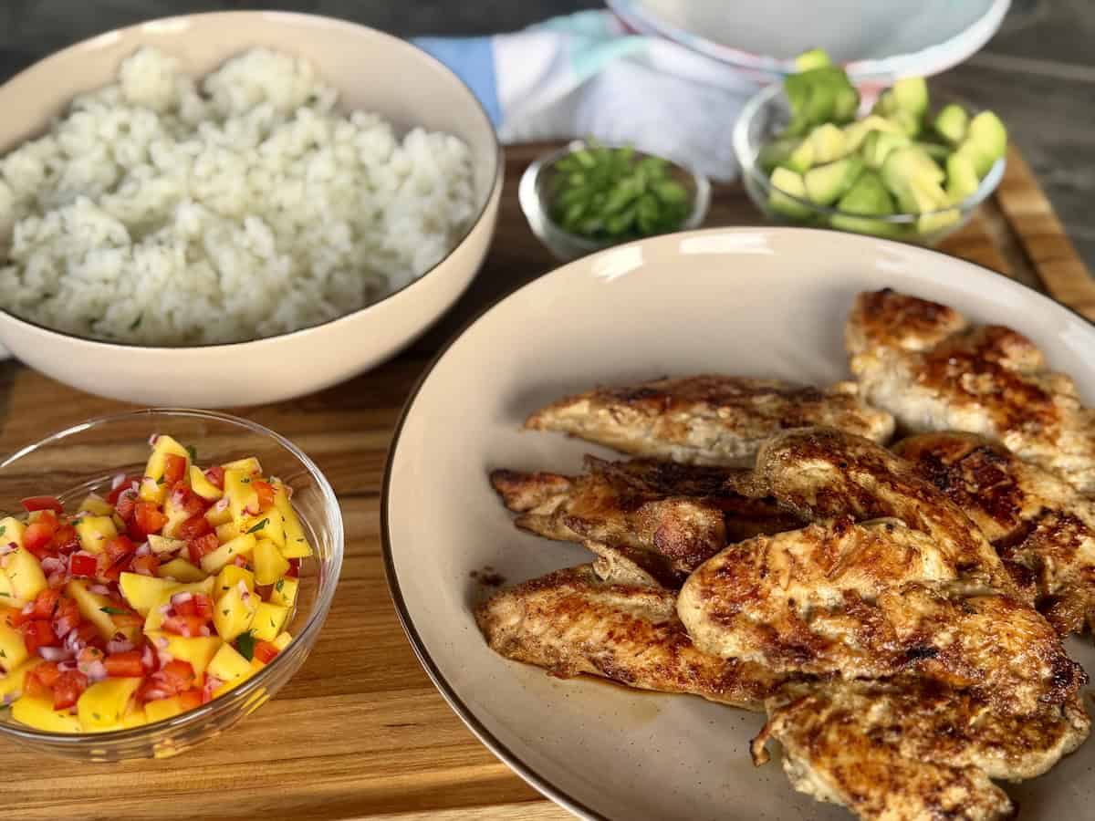 cooked jerk chicken cilantro lime rice mango salsa and diced avocado on wood cutting board to assemble rice bowls