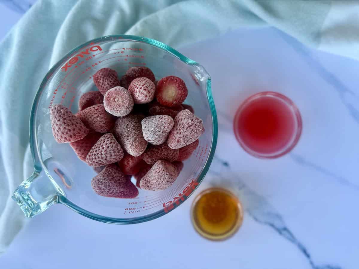 glass bowl of frozen strawberries fruit juice and honey green kitchen towel marble countertop