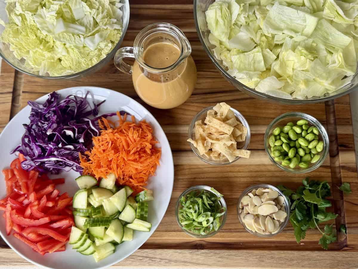 bowls of lettuce cut vegetables and toppings for salad on wood cutting board