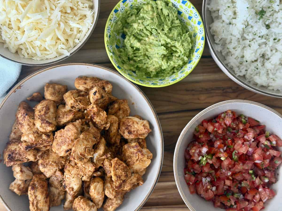 chipotle chicken shredded cheese guacamole cilantro lime rice and homemade pico de Gallo in servings bowls on wood cutting board
